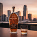 Angels Envy bourbon bottle with a glass of whiskey on a table with a city skyline in the background