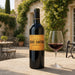A bottle of Château Brane Cantenac Margaux red wine stands on a stone patio next to a glass of red wine, with a blurred stone building and greenery in the background.