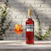 Campari bottle with a cocktail on a wooden surface against a white brick wall.