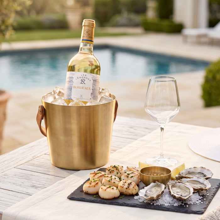 A bottle of Les Legendes Bordeaux white wine in a gold ice bucket on a patio table beside a wine glass, with scallops and oysters on a serving platter in front, set by a swimming pool.