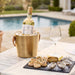 A bottle of Les Legendes Bordeaux white wine in a gold ice bucket on a patio table beside a wine glass, with scallops and oysters on a serving platter in front, set by a swimming pool.