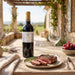 A bottle of Les Legendes Bordeaux red wine stands on a wooden table beside a plate with sliced steak, a wine glass, and a bowl of red grapes, set outdoors on a sunny patio with a vineyard in the background.