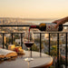 Person pouring red wine into a glass on a balcony with a cityscape view during sunset.