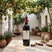 A bottle of Oumouyat Jaspe red wine stands on a stone table beside a filled wine glass, with a cork and corkscrew nearby. The setting is an outdoor courtyard with white walls, hanging grapevines, and potted plants under leafy vines.