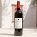 A bottle of Oumsiyat Jaspe red wine stands on a sunlit stone surface against a white wall with soft shadows and a small wooden wall-mounted frame behind it.