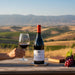 Bottle of Oumsiyat wine with a glass and grapes on a wooden table with a scenic background
