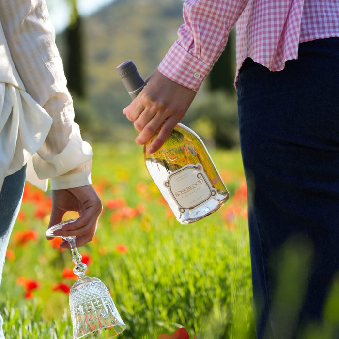 Two people holding a bottle of wine and a glass in a field with flowers and greenery.
