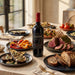 A bottle of Cline Cellars wine stands on a dining table surrounded by plated food, including sliced roast beef, bread, stuffed peppers, and salads, in a warm, sunlit indoor setting.