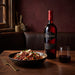 A bottle of Cline Claret Red Zinfandel wine stands on a wooden table beside a bowl of pasta and a glass of red wine, set in a warm, rustic dining room.
