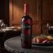 A bottle of Cline Old Vine Zinfandel Red California stands on a dark stone table in a restaurant setting, with a plated steak and garlic in the softly blurred background.