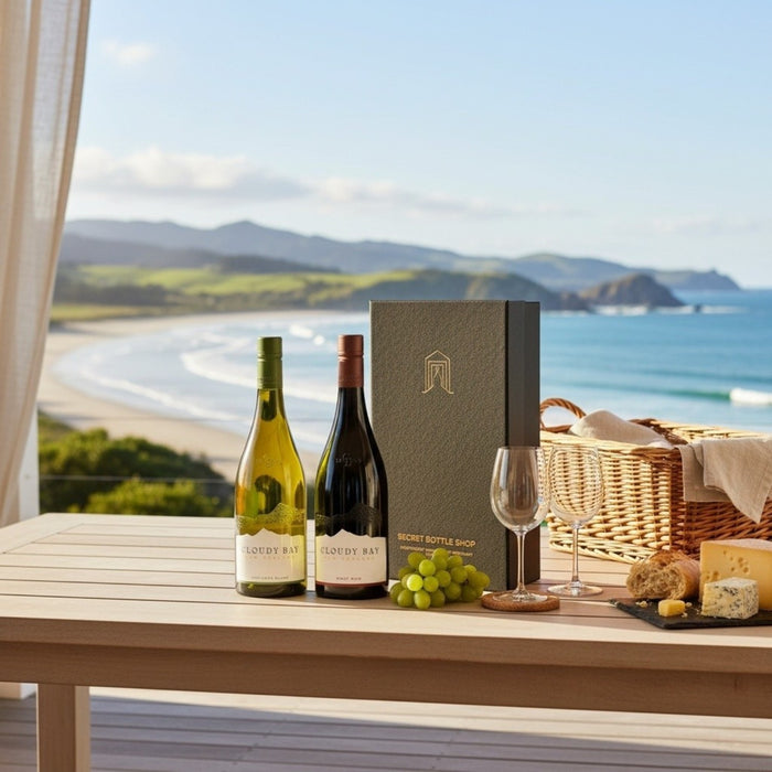 Two wine bottles, a box, grapes, a glass, and a basket on a table with a scenic ocean view.