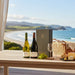 Two wine bottles, a box, grapes, a glass, and a basket on a table with a scenic ocean view.