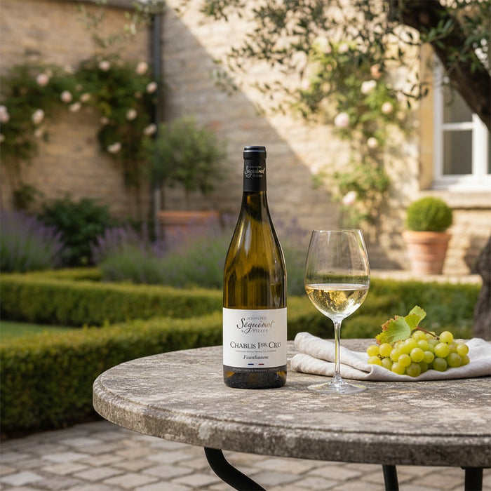 Bottle of Domaine Daniel Seguinot Chablis 1er Cru Fourchaume AOP Chablis with a glass and grapes on a table outdoors.