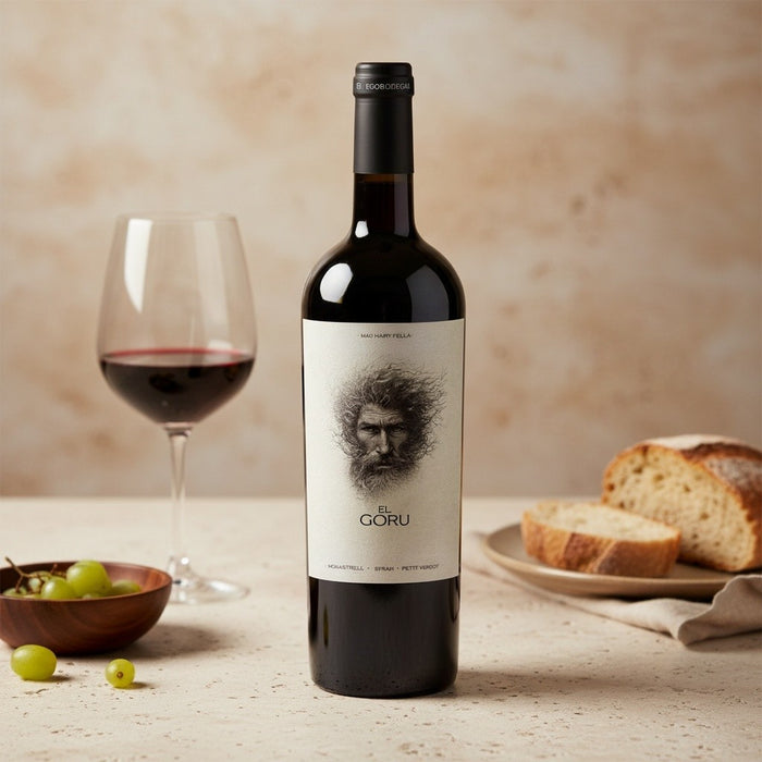 Wine bottle with a label featuring a face, next to a glass of red wine and bread on a wooden table.