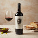 Wine bottle with a label featuring a face, next to a glass of red wine and bread on a wooden table.