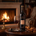 A bottle of Gonzalez Byass Nectar Sherry with a glass of red wine, a lit candle, and wine accessories arranged on a wooden table in front of a cozy fireplace.