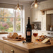 A bottle of Gonzalez Byass Nectar Sherry and glass sit on a wooden kitchen island, surrounded by sliced bread, cheese, nuts, and a corkscrew. A real kitchen interior with cabinets, pendant lights, and a window is visible in the background.