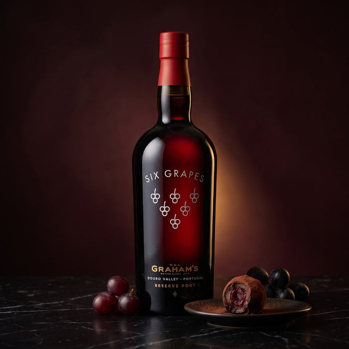 A bottle of Graham's Six Grapes Reserve Port on a dark marble surface, styled with fresh red grapes and a chocolate coated with port wine, against a dark brown background.
