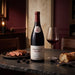 A bottle of Beaujolais Villages red wine by Louis Lator stands on a dark stone table beside a filled wine glass, with sliced steak, blackberries, and a loaf of bread in a warmly lit dining setting.