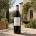 A bottle of Le Mortelle Ampio red wine stands on a stone table beside a filled wine glass, with an outdoor garden and building in the background.