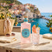 A bottle of Malfy Gin Gin sits on a marble table in front of a coastal village and blue sea, styled with a cocktail glass, sunglasses, a woven tote bag, and lemon branches.