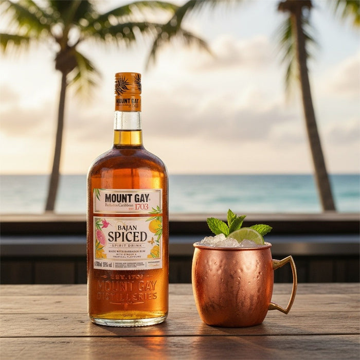 Mount Gay Spiced rum bottle with a copper mug of cocktail on a wooden table by the ocean.