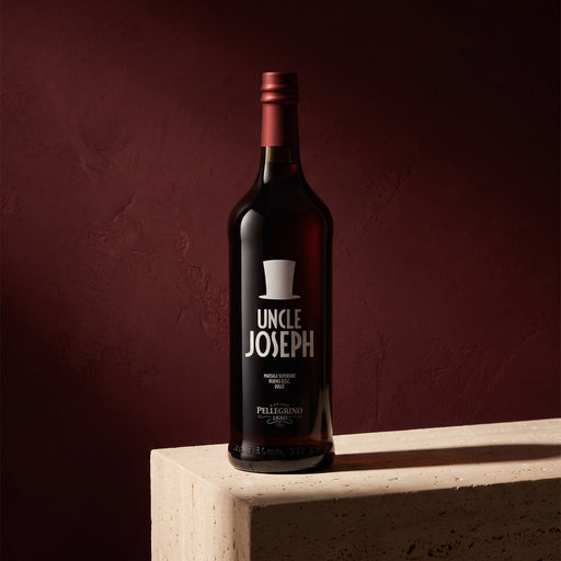 A bottle of Uncle Joseph Marsala  stands on a light beige stone pedestal against a dark, textured burgundy background. The bottle is dark glass with a metallic red foil top and a black label featuring a white top hat logo and the brand name 'UNCLE JOSEPH'.