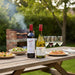 Two wine bottles on a wooden table outdoors with food and glasses.
