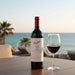 Penfolds wine bottle with a glass of red wine on a table by the ocean.