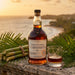 A bottle of Four Square Rum Distillery 14 Year Old rum and a matching glass on a stone ledge, surrounded by green palm leaves and a bundle of sugarcane, with a beach sunset in the background.