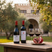 Two bottles of wine with two glasses and a pomegranate on a wooden table outdoors.