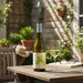 Bottle of white wine and a glass on a wooden table outdoors with plants and furniture in the background.