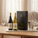 Two wine bottles with a black box on a table with glasses and ice bucket.