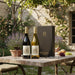 Two wine bottles and a gift box on a table outdoors with a stone building and flowers in the background.