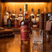 A bottle of Weller Antique 107 whisky stands on a polished wooden bar counter next to a cut crystal whisky glass, with a blurred background of other bottles and books.