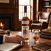 Bottle of Willett bourbon on a wooden table with a glass and a hand holding a pen, in a cozy room with a fireplace and books.