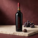 A bottle of Malbec wine with a black label and red script logo stands on a stone slab next to a cluster of dark grapes and a chocolate, against a textured dark red-brown background.