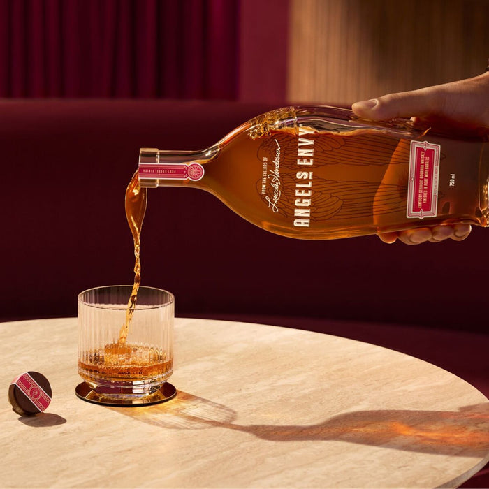 Angels Envy bourbon being poured into a glass on a marble surface with a red curtain background.