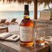 A bottle of Foursquare Epilogue single blended rum sits on a wooden table at a beachside bar, with a filled rocks glass, an open book, and lounge chairs in the background.