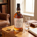 A bottle of Foursquare Epilogue Rum stands on a wooden table in a warm, sunlit room. A clear glass, a small dropper bottle, and a leather-bound notebook are arranged beside it, with a bookshelf and window in the background.