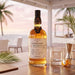 A bottle of Foursquare Epilogue Rum stands on a wooden table in a bright, airy patio setting with a beach and palm trees blurred in the background. A clear whisky glass, a metal jigger, and a lemon twist are placed beside it.