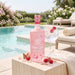A bottle of Rockfield Pink Gin is displayed on a stone table next to a cocktail glass with raspberries, set against a luxurious outdoor poolside background with lounge chairs and greenery.