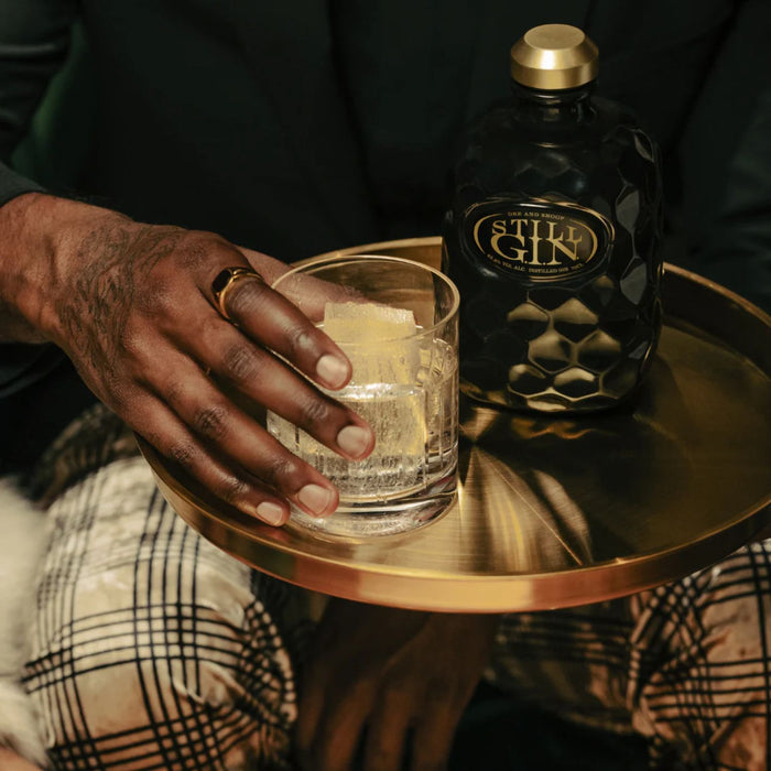 Person holding a glass of gin with a bottle of Still G.I.N. Gin By Dr. Dre & Snoop Dogg on a small round table.